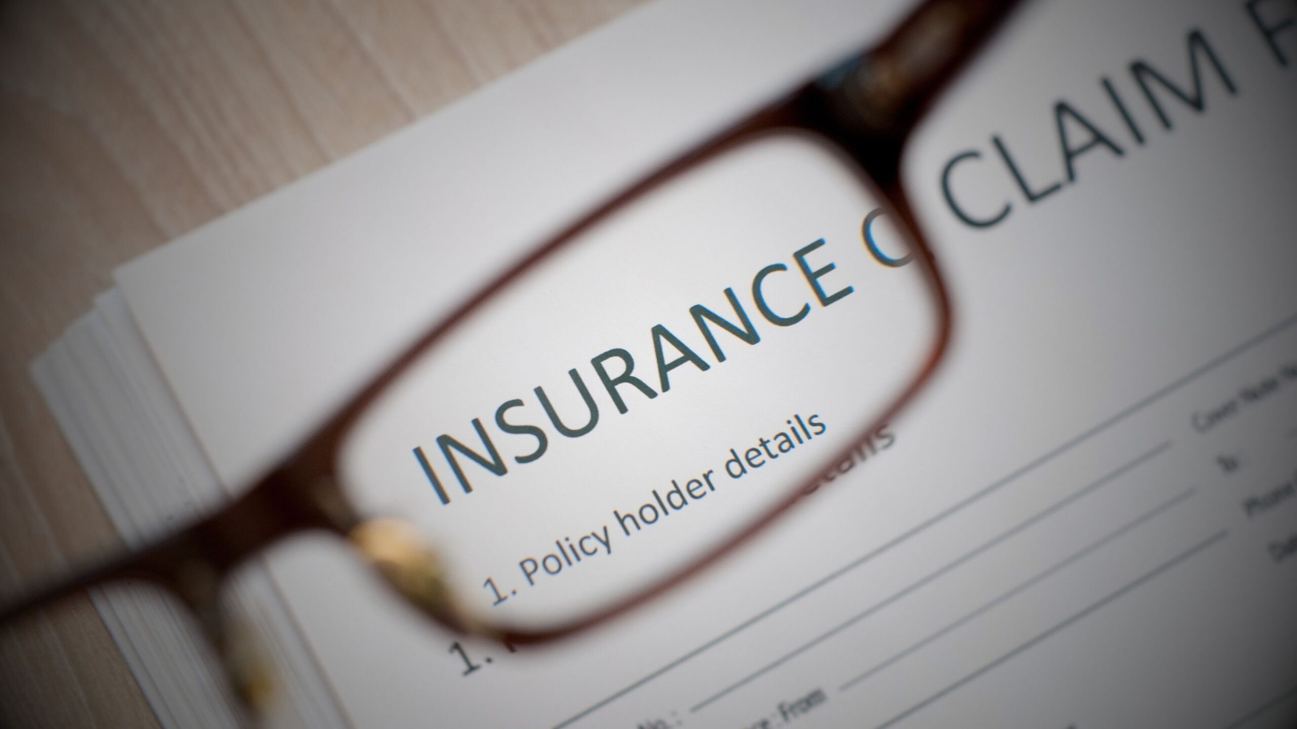 Close-up image of a pair of reading glasses resting on an insurance claim form, focusing on the word "INSURANCE" and "Policy holder details," symbolising claims administration and policy review.