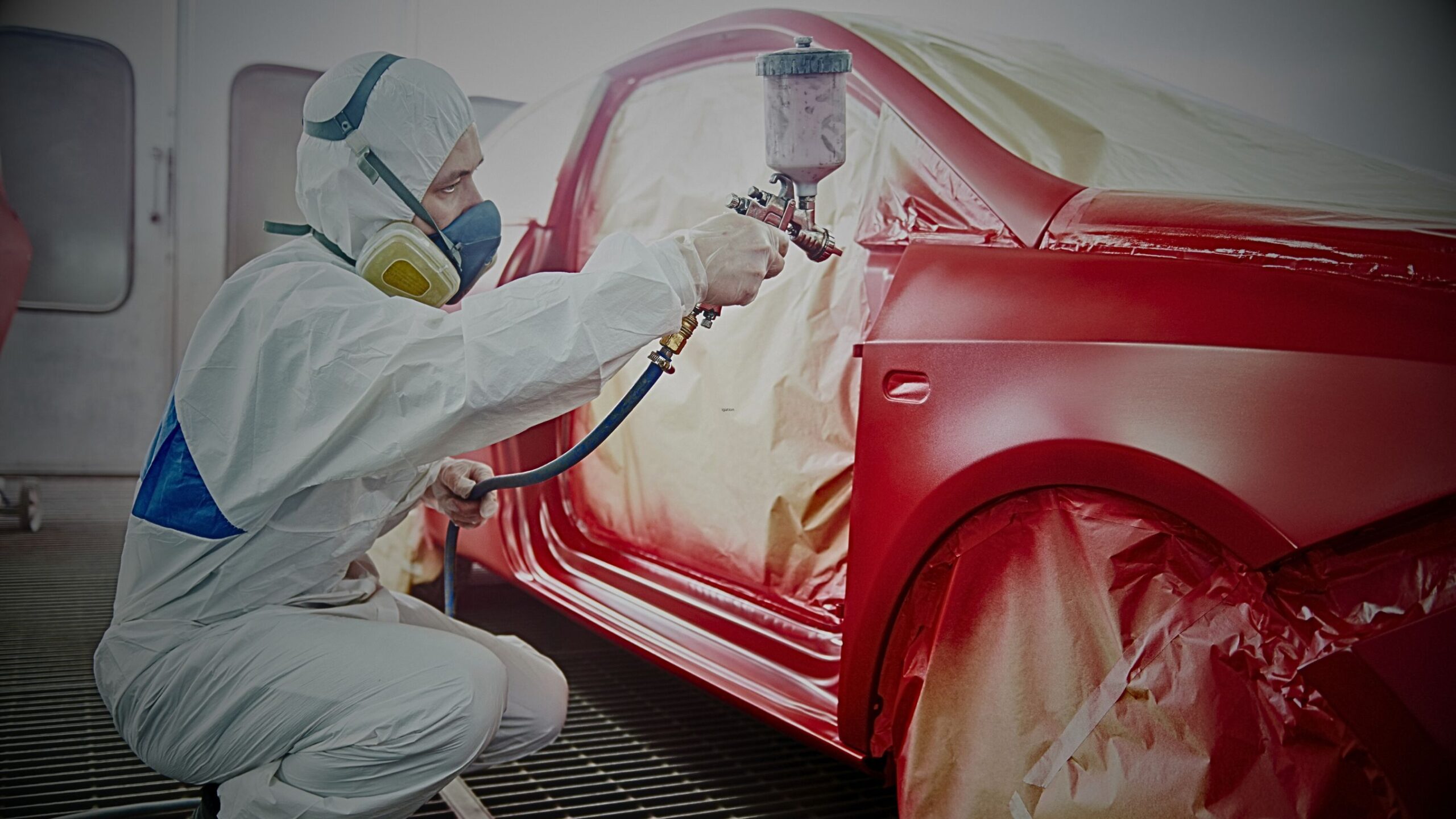 Automotive technician wearing protective gear and respirator, applying a coat of red paint to a car body panel with a spray gun inside a bodyshop, illustrating high-quality vehicle repair and refinishing.