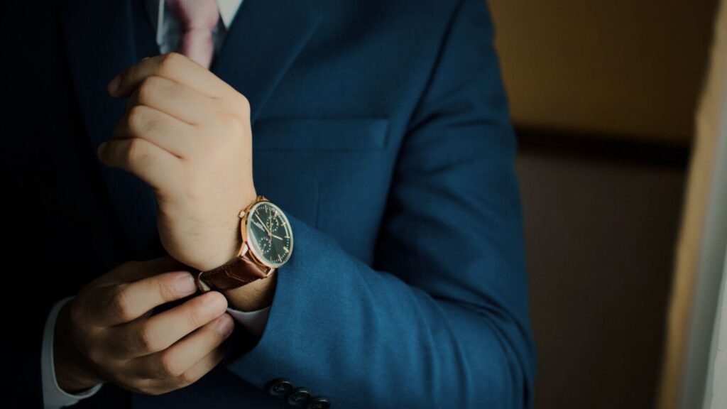 Close-up of a man in a blue suit and pink tie adjusting a luxury timepiece with a brown leather strap, symbolising high-value asset, watch, and jewellery insurance.