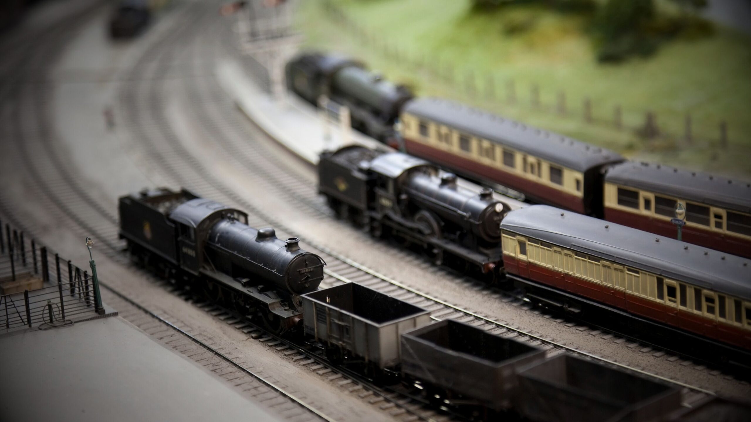 A high-angle, shallow depth-of-field shot of an intricate O-scale or OO-scale model railway layout featuring several detailed black steam locomotives pulling passenger and freight cars on parallel tracks.