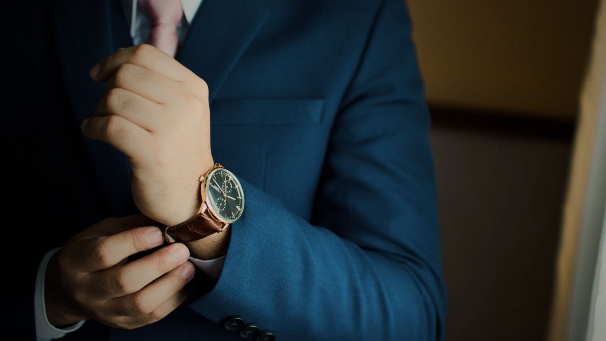 Close-up of a man in a blue suit and pink tie adjusting a luxury timepiece with a brown leather strap, symbolising high-value asset, watch, and jewellery insurance.