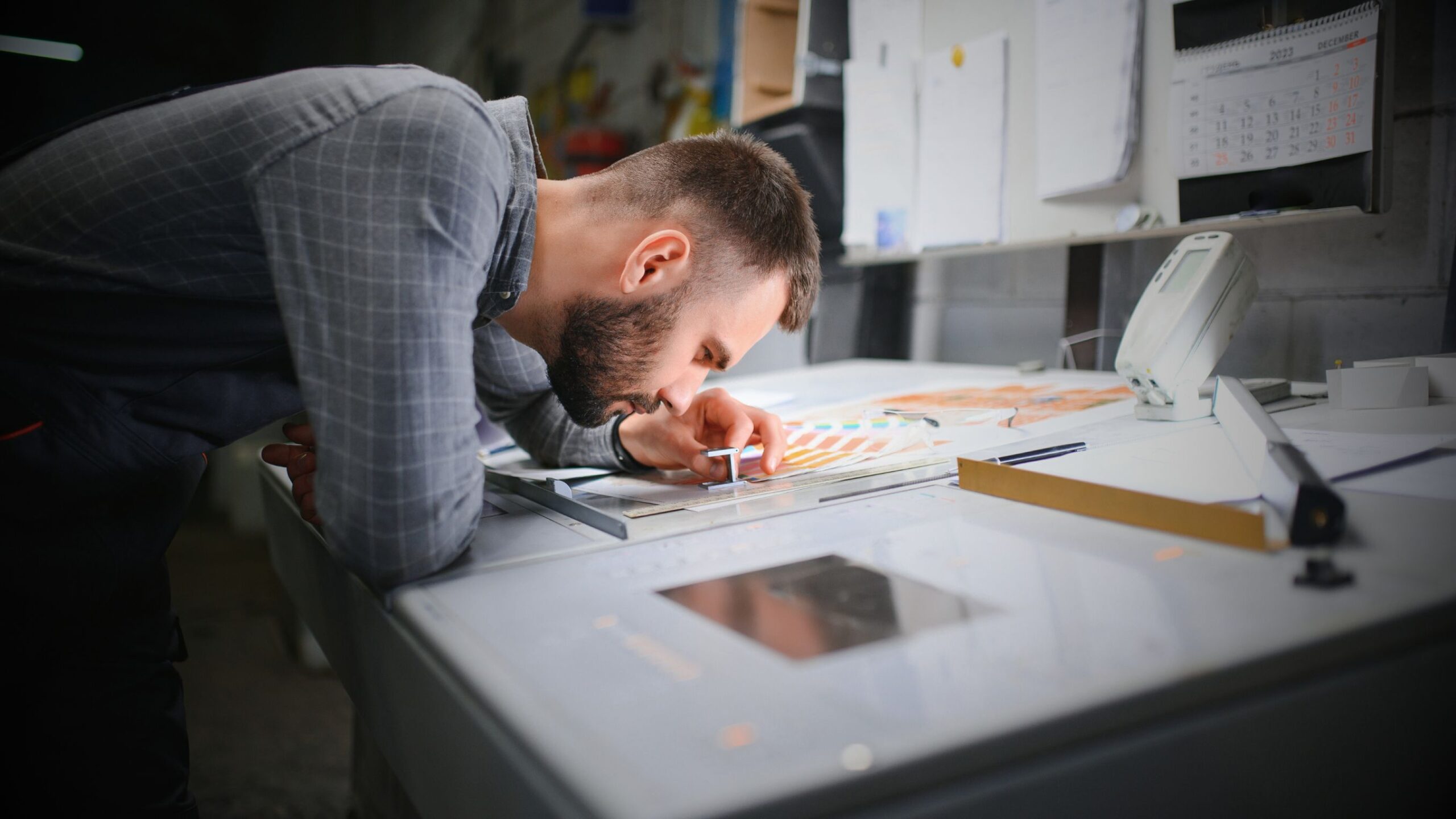 Printer Employee A bearded man, appearing to be a designer, printer, or quality control technician, is leaning over a large workbench, closely examining colour swatches and plans with a precision tool. The scene suggests technical printing, design, or manufacturing work.