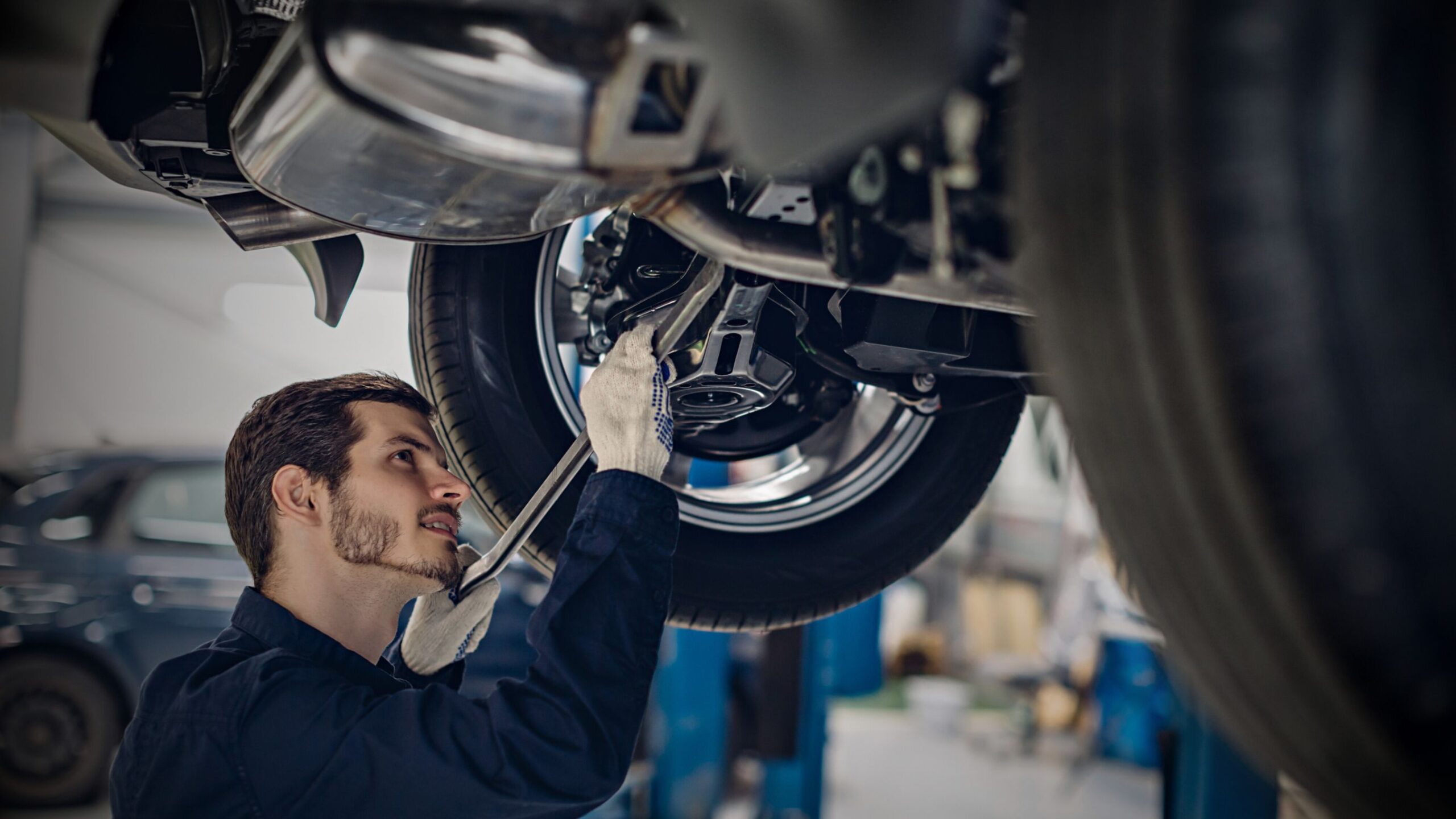 Motor Technician A smiling vehicle technician with a beard, wearing work gloves, is working underneath a raised vehicle, inspecting or repairing the suspension and wheel assembly in a well-lit professional garage or automotive workshop.