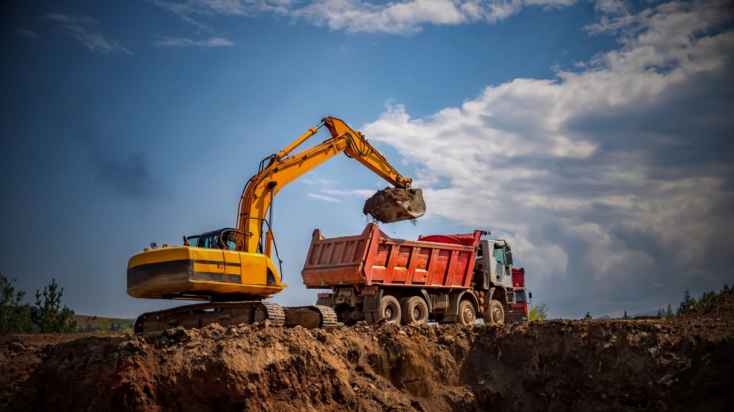 Industrial Business Heavy construction site showing a yellow tracked excavator loading soil/dirt into the back of a red dump truck under a bright, cloudy sky. Image highlights the need for construction and plant insurance.