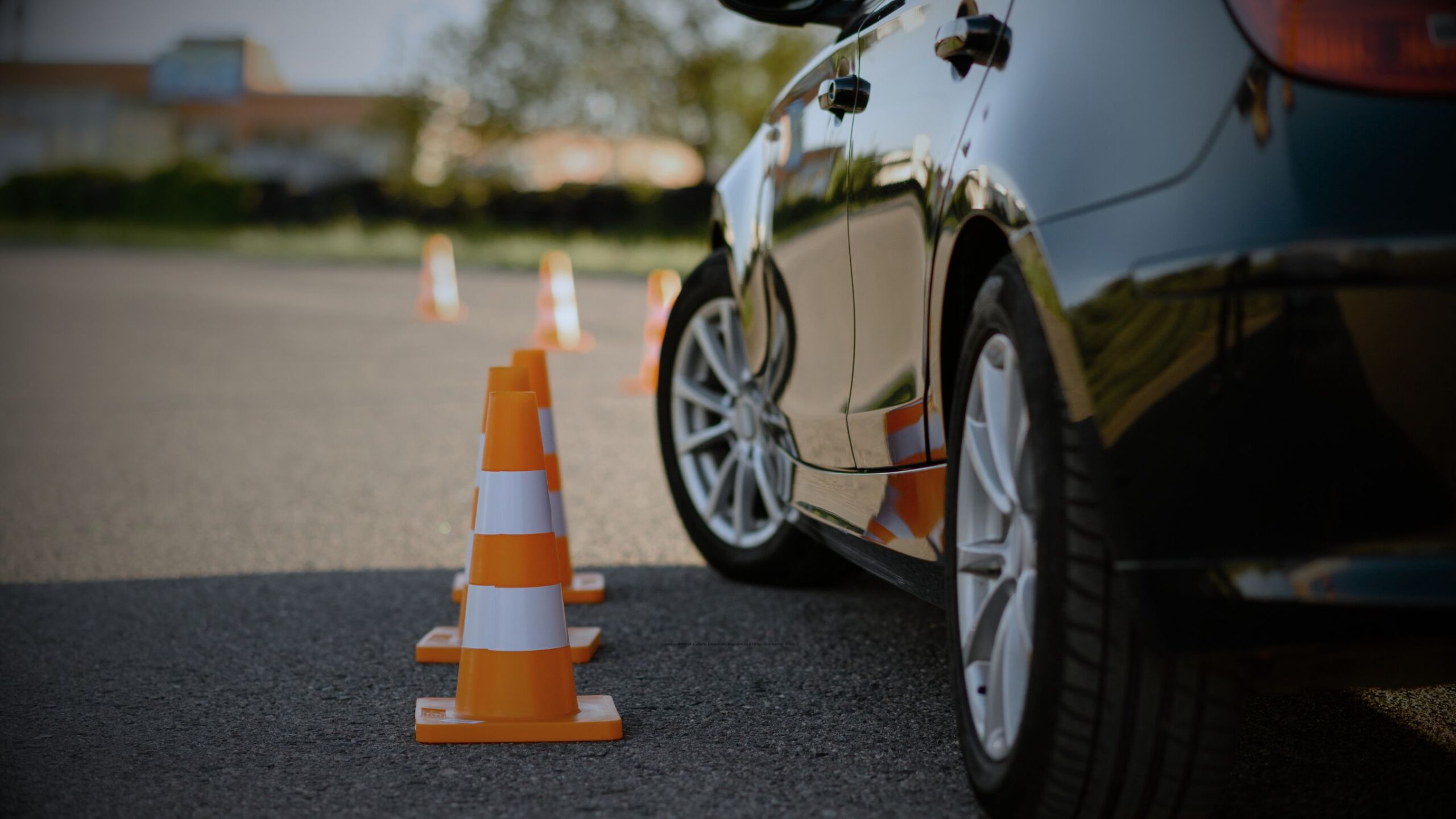 Low-angle close-up of the rear wheel of a dark car passing near a line of orange and white traffic cones, symbolising driving practice and manoeuvres for learner drivers, promoting VINTTRO insurance.