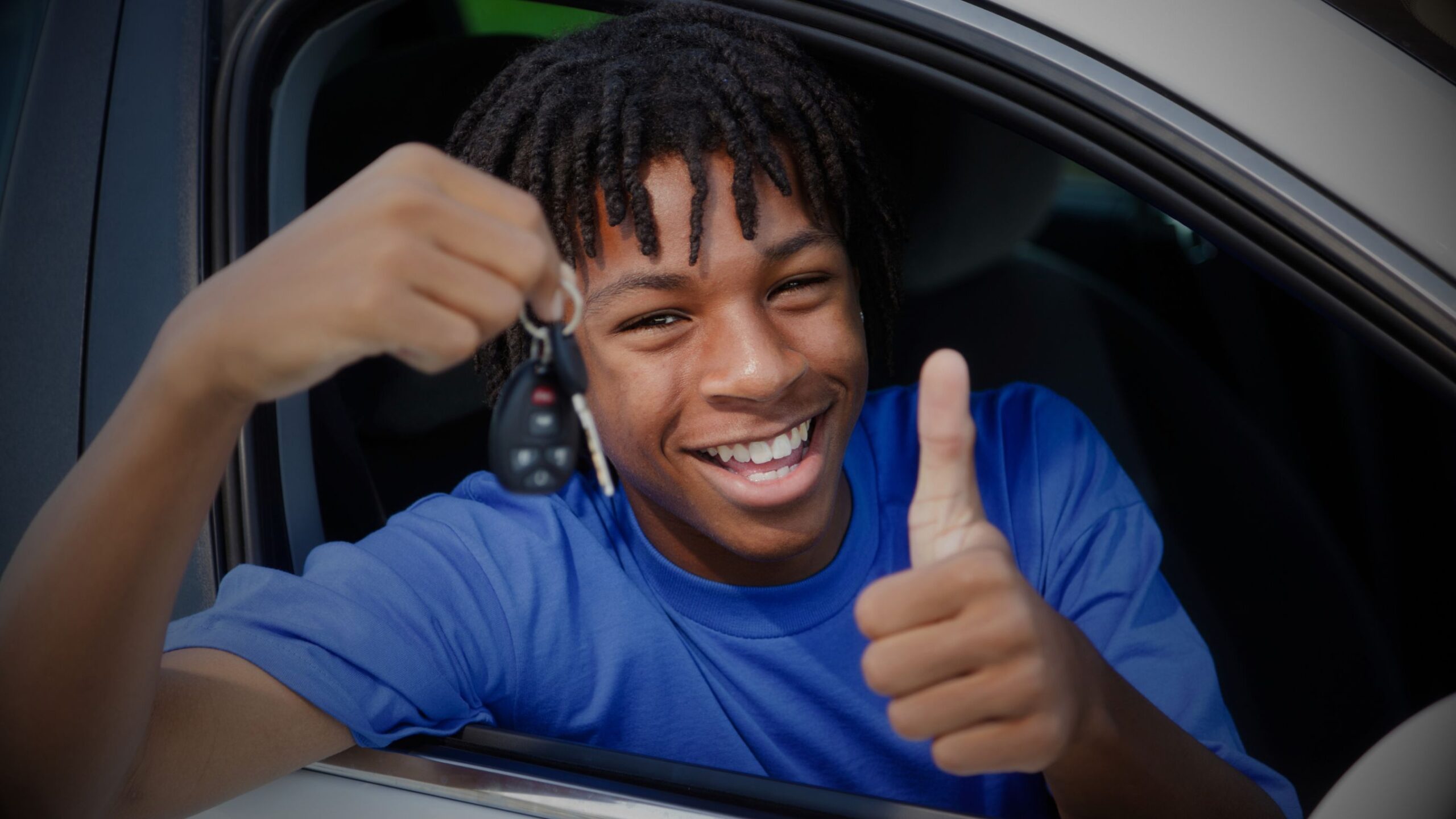 Smiling young person in a blue shirt giving a thumbs-up and holding car keys through the window of a car, representing the excitement of learning to drive with VINTTRO Cover's learner driver insurance.