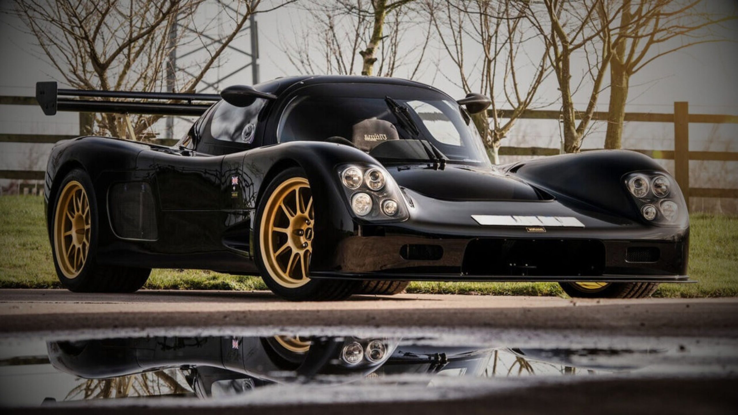 Kit Car Black, low-slung performance kit car, resembling an Ultima GTR, with gold wheels and a large racing wing, reflected in a puddle. VINTTRO Cover high-performance kit car insurance.