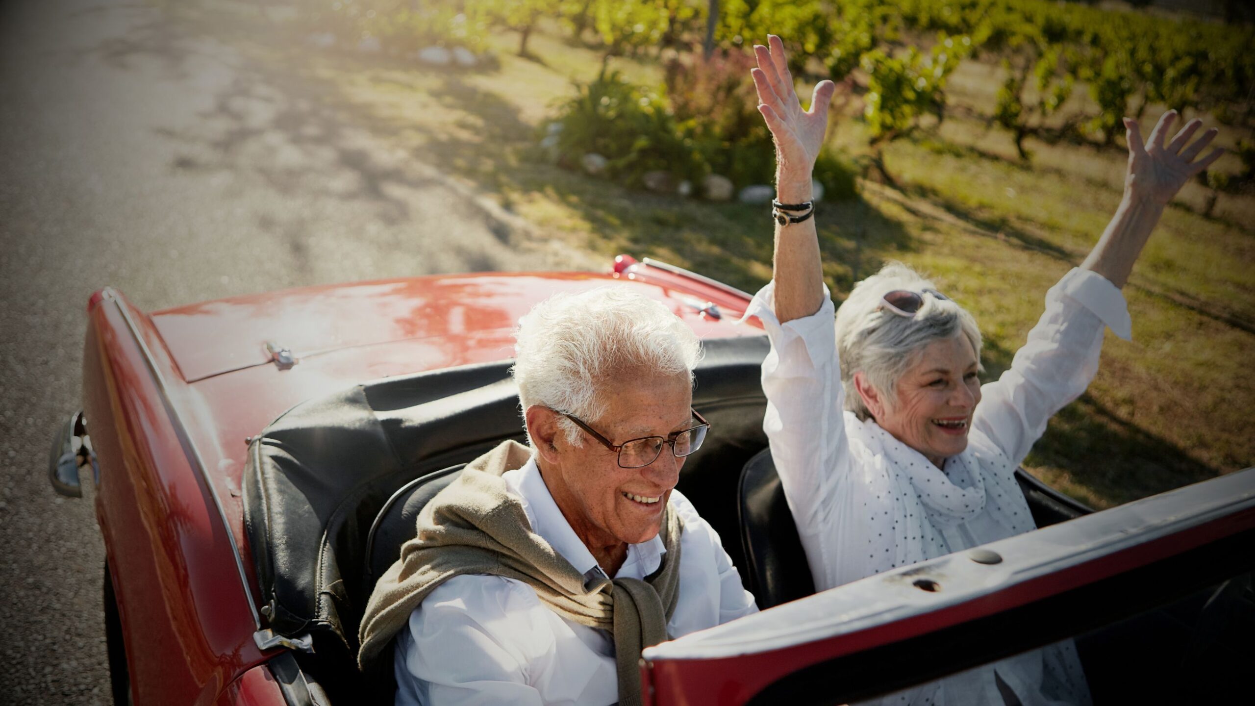 A joyful, older couple driving a red classic convertible car with the woman raising her arms in excitement, symbolising financial freedom and security in retirement, supported by VINTTRO's life funding and protection insurance.