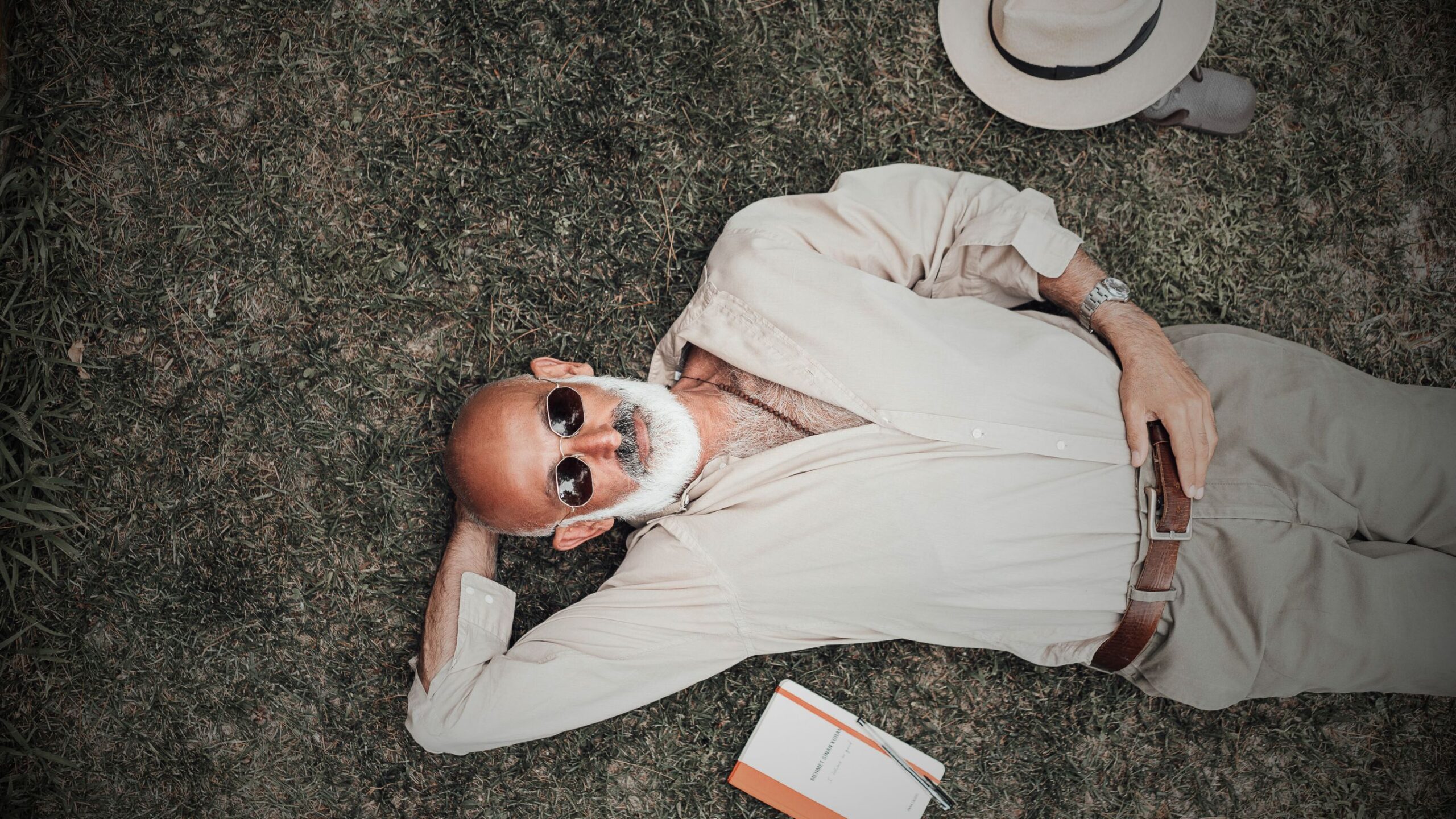 Mortgage and Protection, this image displays an overhead view of a stylish man with a white beard and sunglasses relaxing on the grass next to a notebook and a hat, symbolising guaranteed income protection and financial freedom secured by VINTTRO's financial solutions.