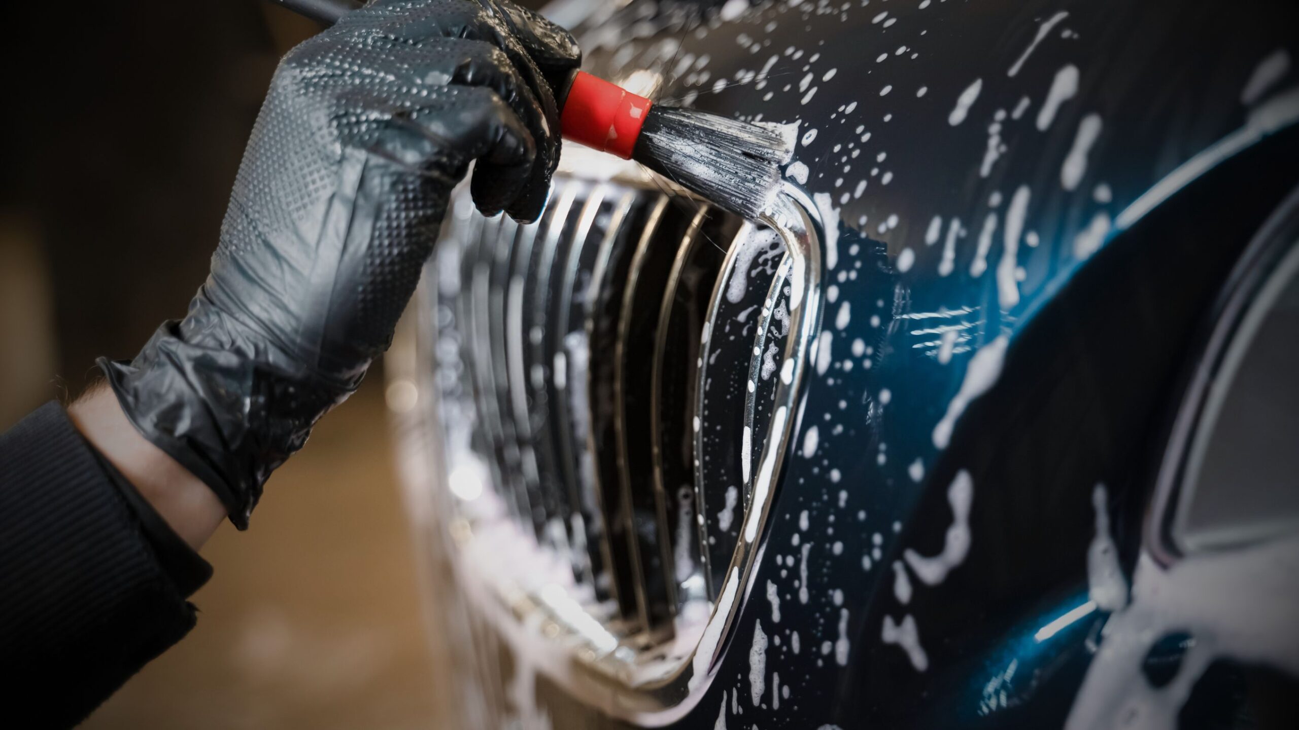 Valeting & Detailing Close-up of a black-gloved hand cleaning a car grille with a detailing brush and soap foam.
