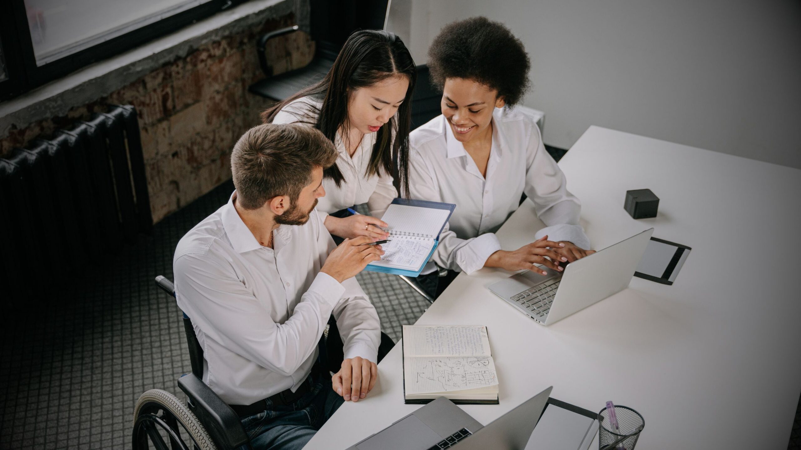 Happy Workforce Three diverse VINTTRO B2B professionals, including one using a wheelchair, collaborating on a project with a laptop and notebook, symbolising inclusive teamwork and strategic business consulting.