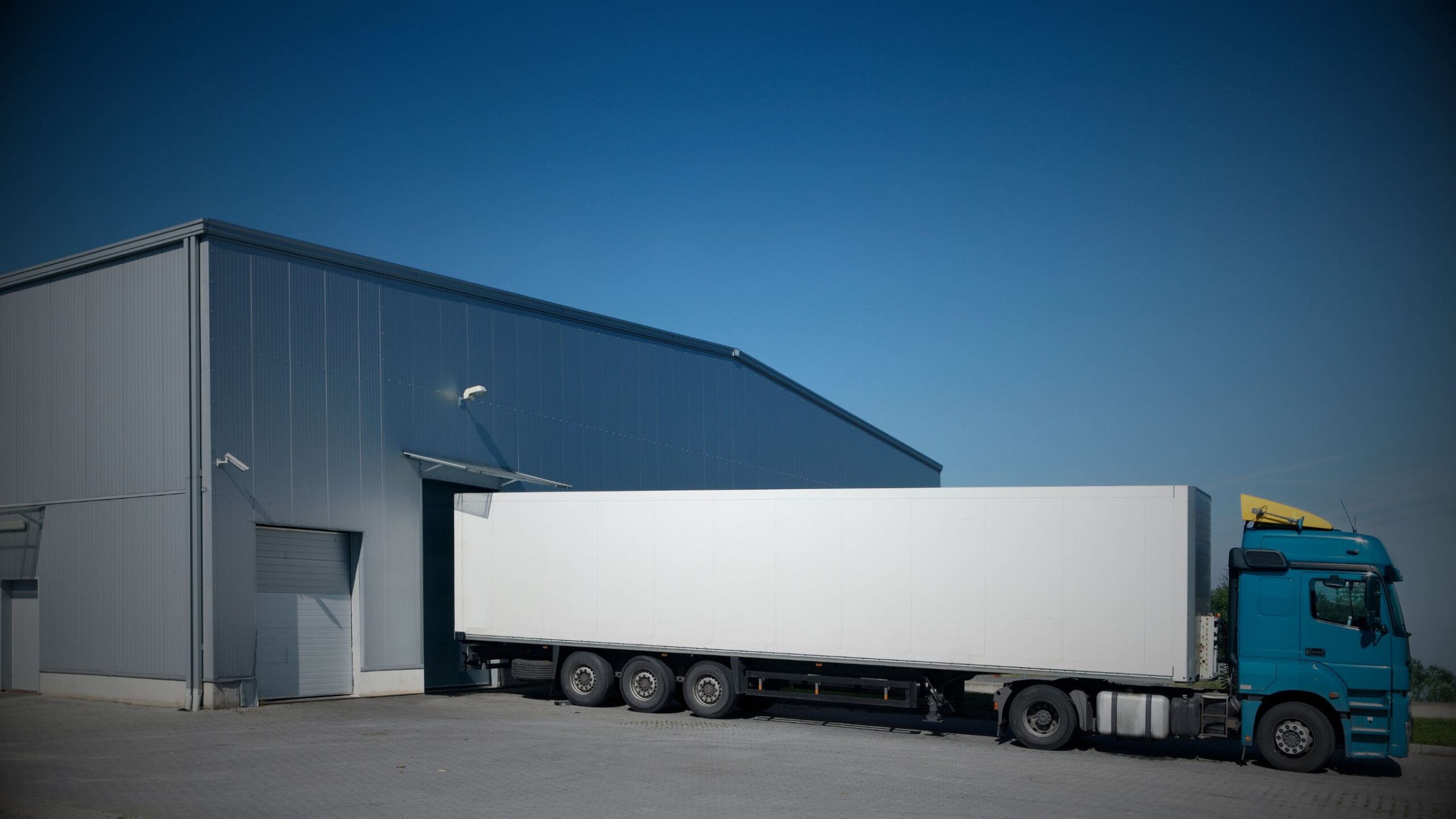 Semi-truck with a white trailer backed into a loading bay of a modern grey warehouse/distribution centre, representing logistics, transport, and commercial contract legal services.