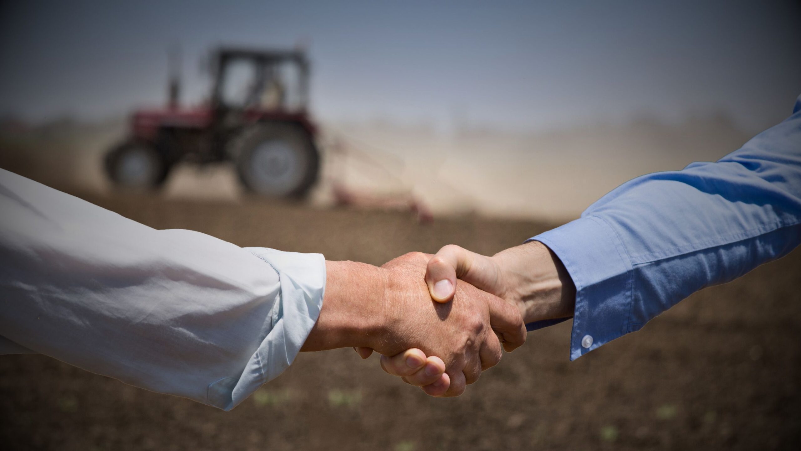 Conveyancing Legal Services Business handshake in a large agricultural field with a tractor, symbolising completion of farm land conveyancing deal in the UK.