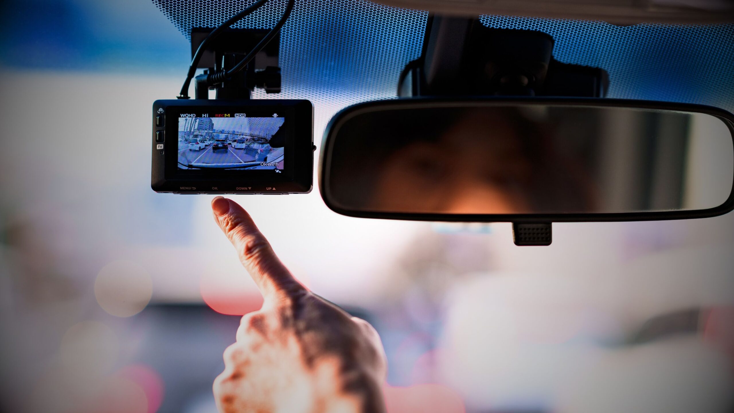 Driver's hand pointing at a dash camera screen mounted on a windshield, with a rear-view mirror visible, symbolising dash cam installation and telematics technology.