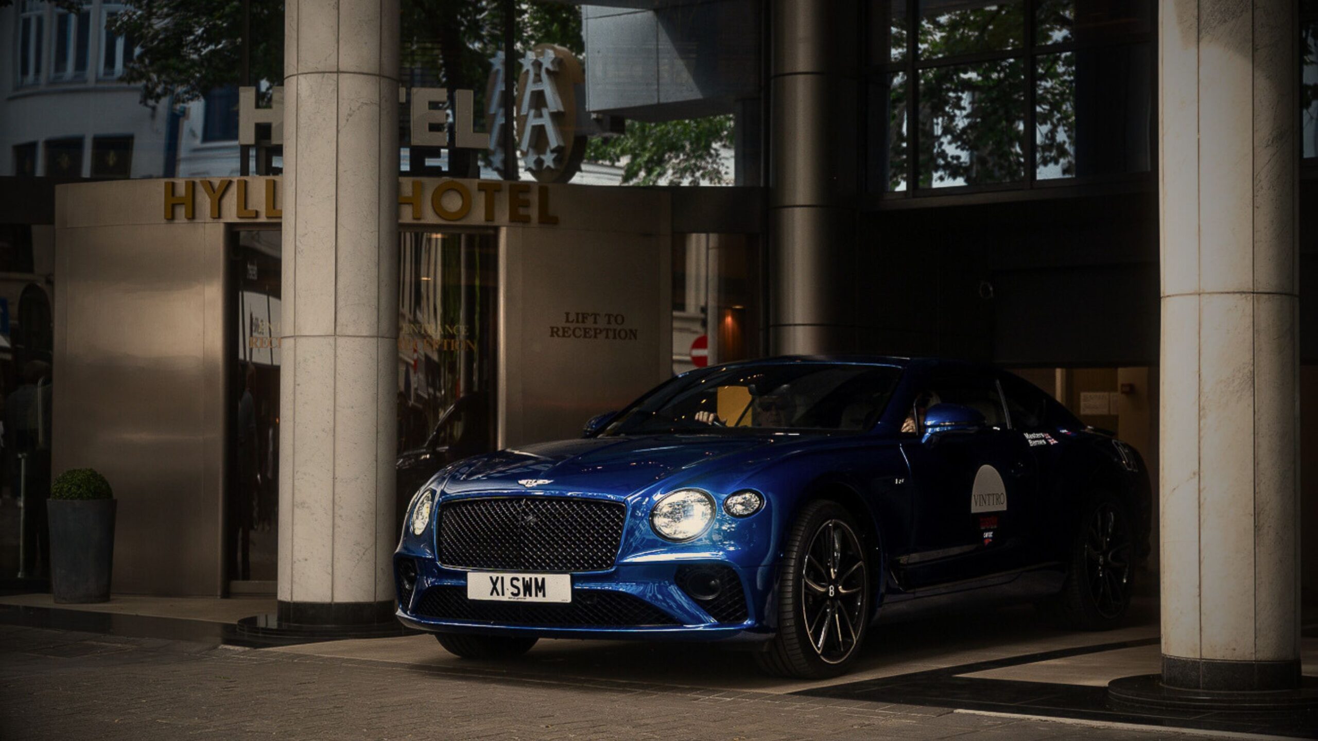 A royal blue Bentley Continental GT with VINTTRO branding exiting the Hyatt Regency Hotel during a luxury automotive networking event.