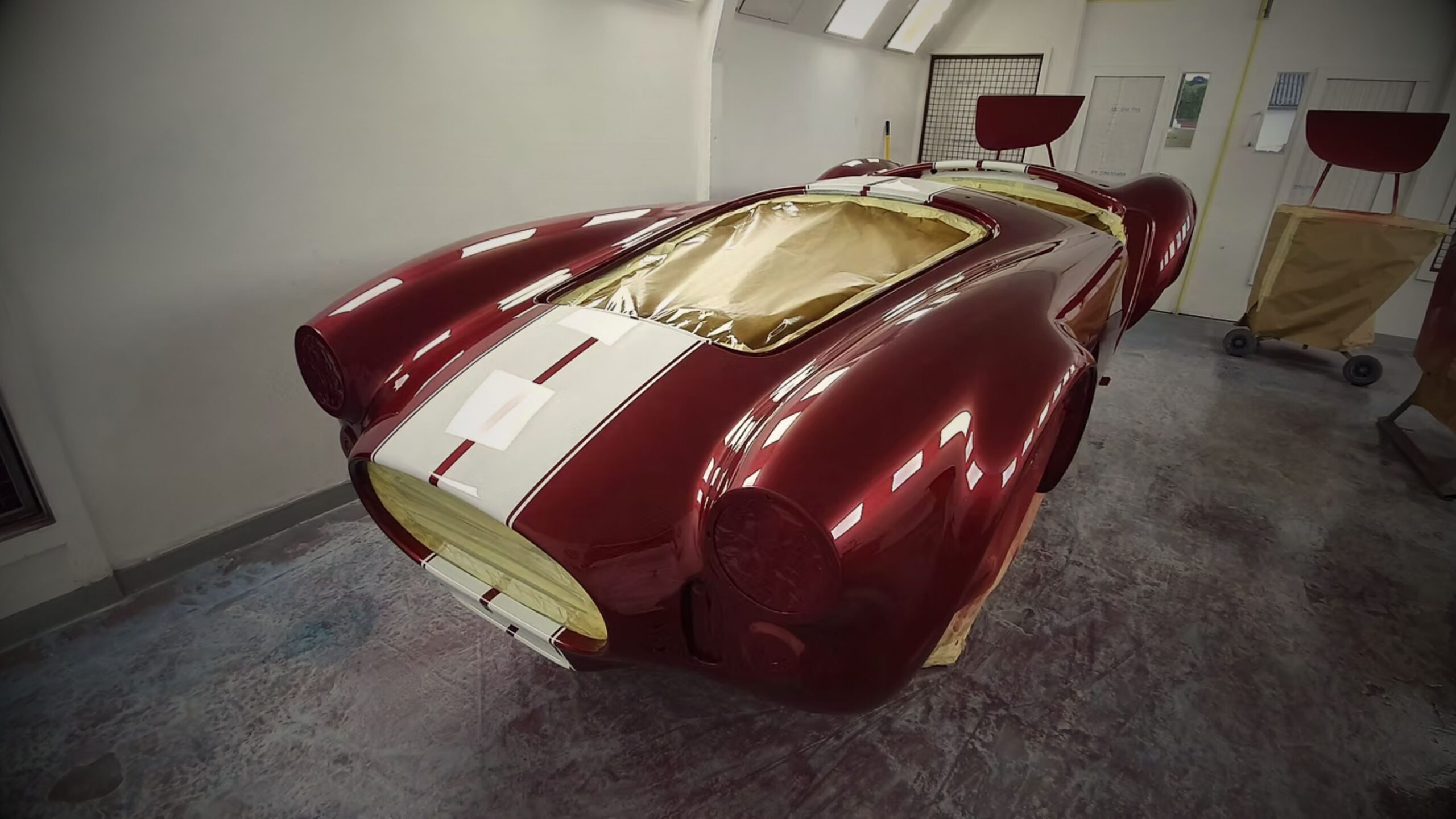 Repair and Restorations Services showing a classic AC Cobra body shell undergoing a professional candy apple red respray with white racing stripes in a specialist automotive paint booth.
