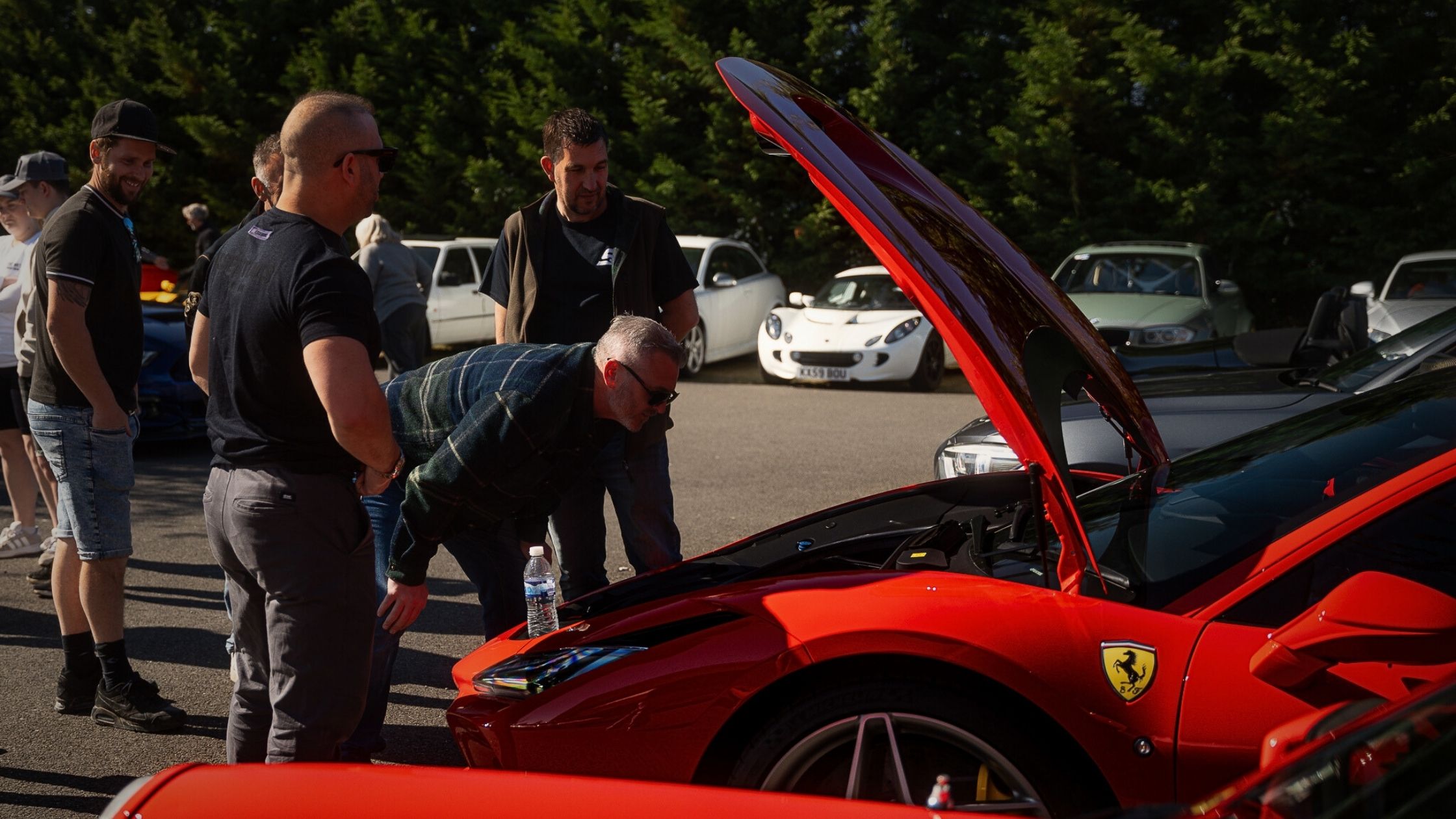 A group of car enthusiasts gathered in a sunlit car park, leaning in to inspect the engine bay of a bright red Ferrari with its bonnet open at an automotive community event.