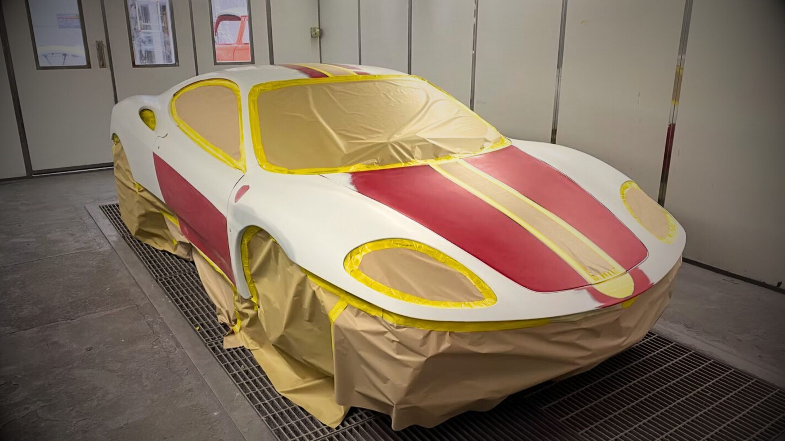 A Ferrari 360 Modena in a professional spray booth undergoing a multi-stage respray, meticulously masked with yellow tape and protective paper, showcasing VINTTRO’s specialist supercar paint services.