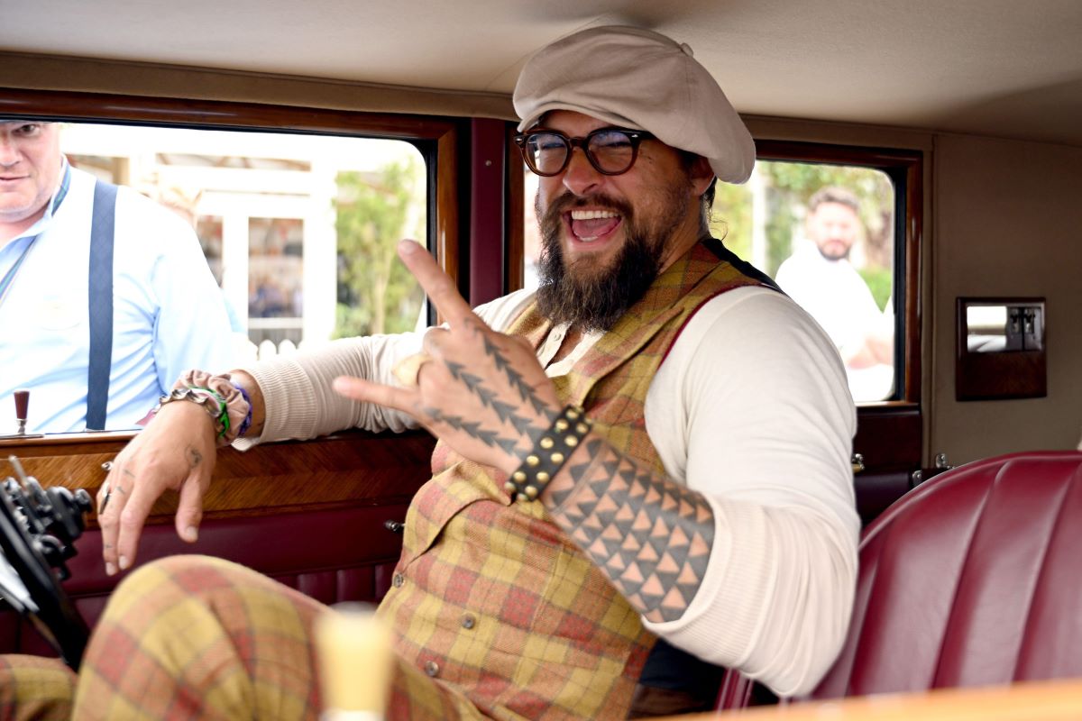 A side profile of Jason Momoa in a 1929 Rolls-Royce Phantom II vintage car in a deep polished finish, featuring its iconic long hood and classic spoked wheels, converted to an electric vehicle by Electrogenic