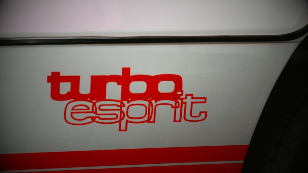 Lotus Esprit: A close-up detail of the iconic red 'turbo esprit' decal on the side of a white classic Lotus Esprit, showcasing the 1980s typography and styling.