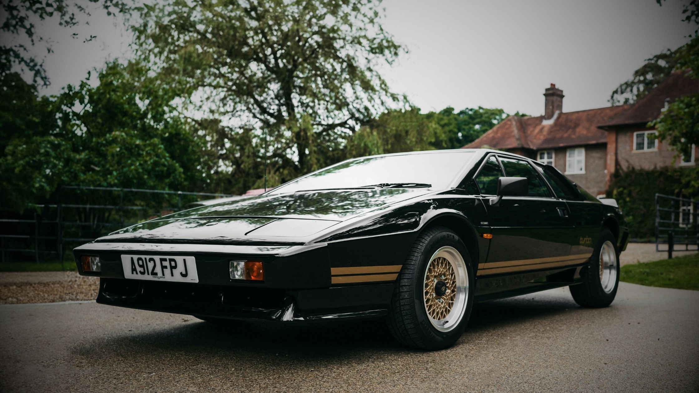 Lotus Esprit Turbo in classic black and gold JPS colours parked on a gravel driveway in front of a traditional British manor house, celebrating 50 years of Lotus heritage.
