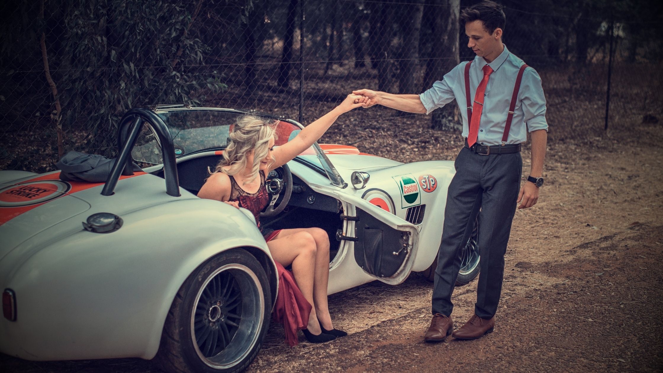 A young man in a white shirt and red suspenders holding the hand of a young woman in a red gown as she exits a silver classic sports car with racing decals.