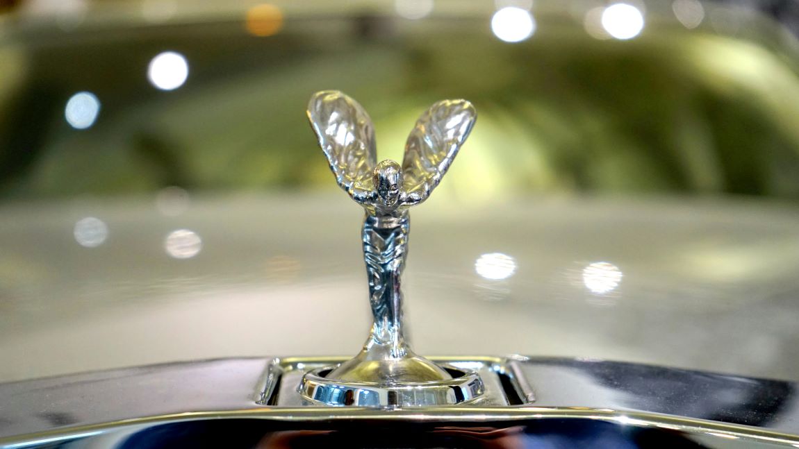 Rolls-Royce: Close-up of the iconic silver Spirit of Ecstasy hood ornament, also known as the Flying Lady, on the polished chrome grille of a luxury Rolls-Royce.