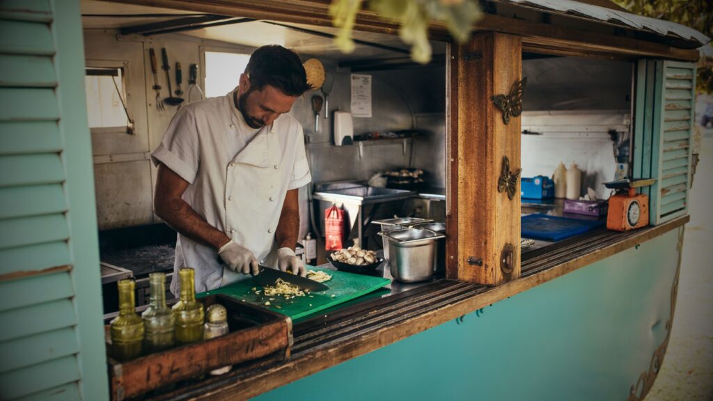 A chef in a white uniform preparing food inside a teal-coloured vintage food trailer, highlighting the professional equipment and custom fit-outs covered by VINTTRO Covers specialist business vehicle insurance.