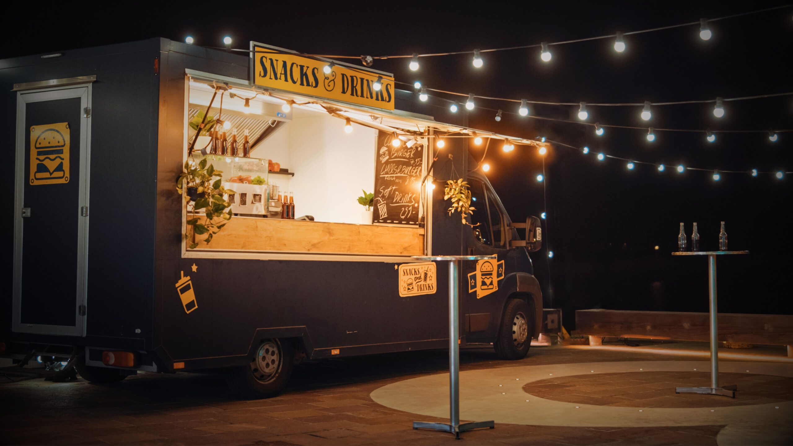 A modern black food truck lit with warm string lights at night, featuring a "Snacks & Drinks" sign and outdoor bistro tables, illustrating a mobile catering business insured through VINTTRO Cover, who provides specialist vehicle insurance.