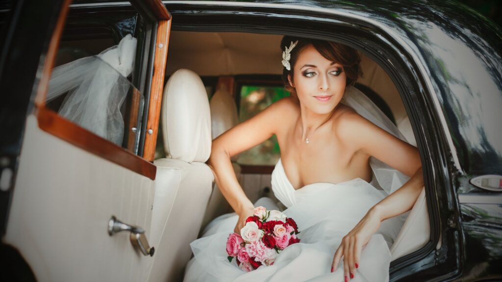 A bride in a white strapless wedding gown and veil sitting in the back of a vintage black luxury car with cream leather interior, holding a bouquet of red and pink roses.