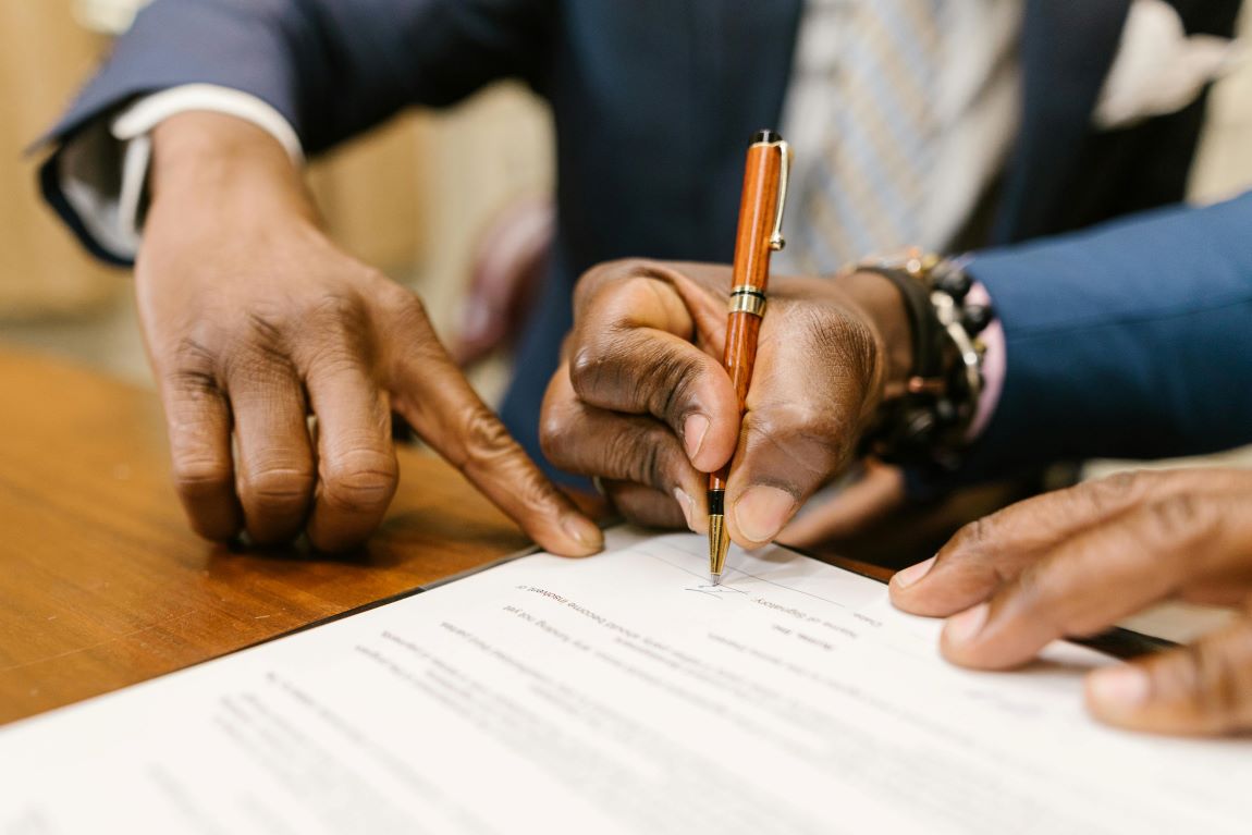 Will Writers. Close-up of a person in a professional suit signing a legal document with a fountain pen while another person points to the signature line on a wooden desk.