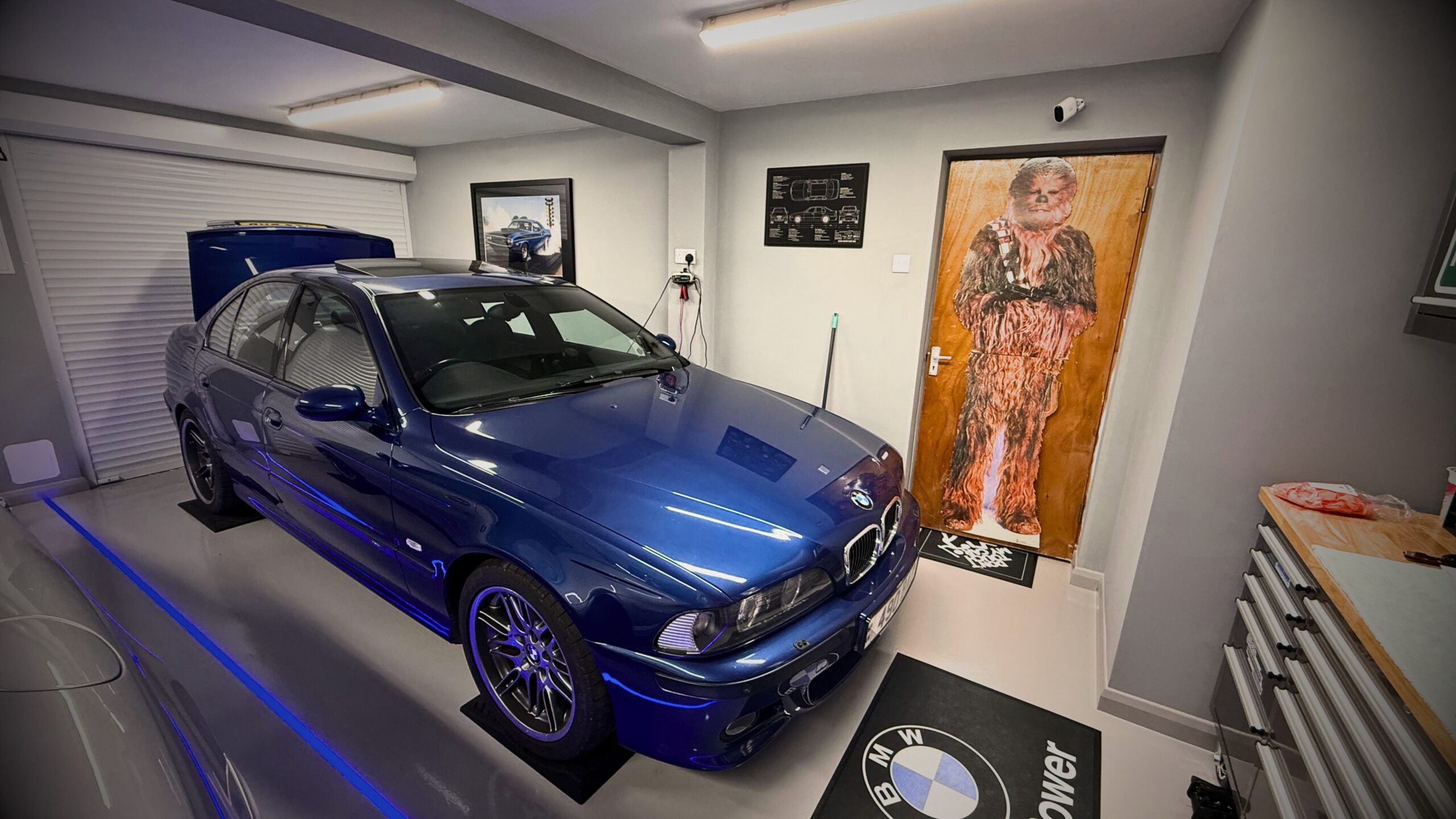 Close-up of VINTTRO members Greg’s immaculate home showroom in the UK, featuring a polished blue BMW E39 M5 on custom M Power mats, with a Chewbacca cardboard cut-out, car schematics, and clean workshop storage visible.