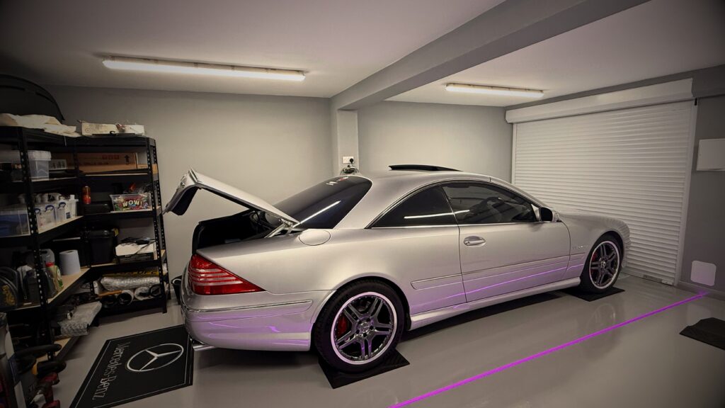 Side profile of a pristine silver Mercedes-Benz CL coupe in a custom UK home showroom, with the boot open, resting on branded Mercedes-Benz mats. The image shows tidy garage storage shelving, a white roller shutter door, and clean grey resin flooring with purple under-car LED lighting.