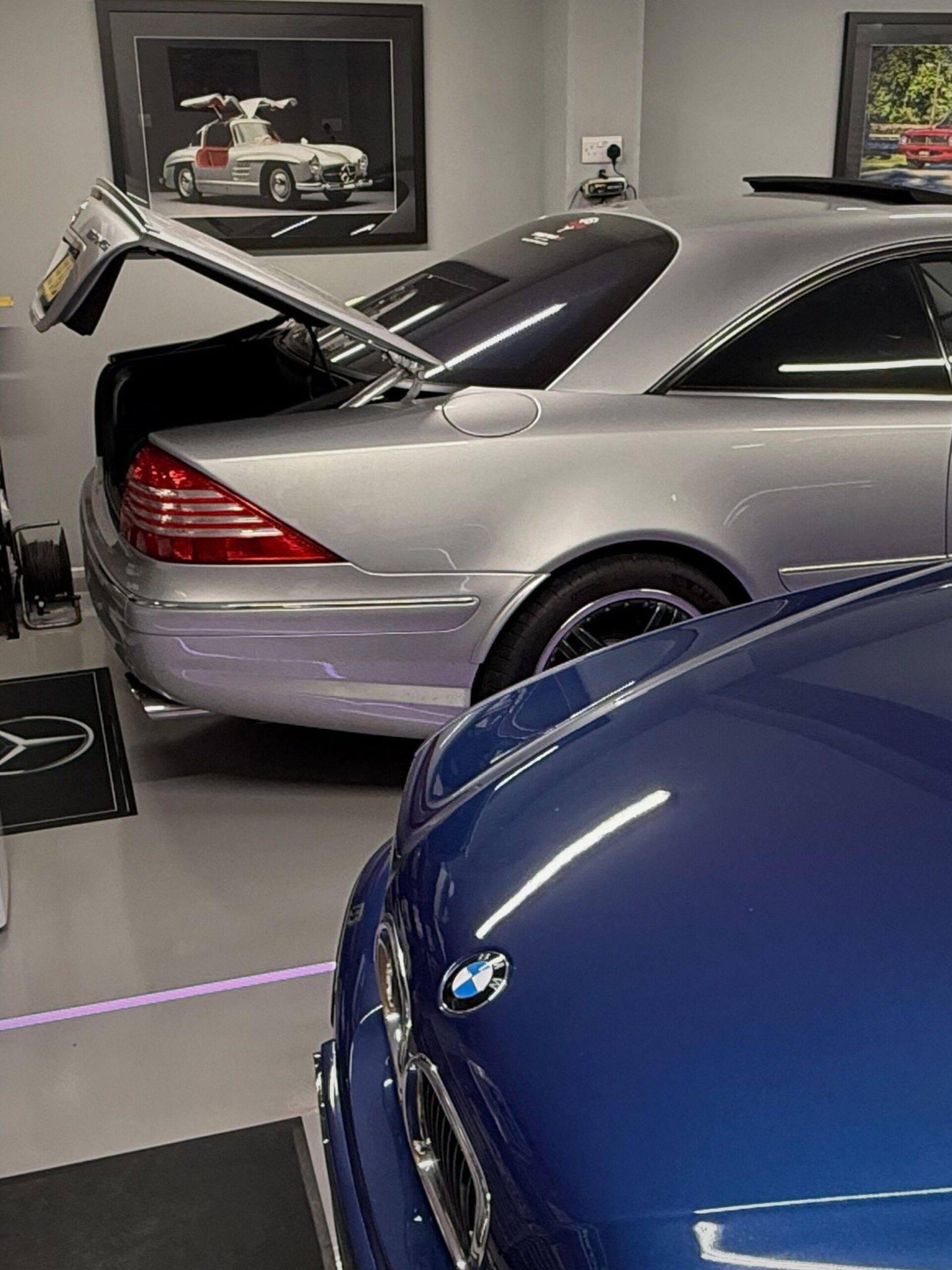Close-up of VINTTRO members Greg’s immaculate home showroom in the UK, featuring a polished blue BMW E39 M5 on custom M Power mats, and Mercedes AMG with a Chewbacca cardboard cut-out, car schematics, and clean workshop storage visible.