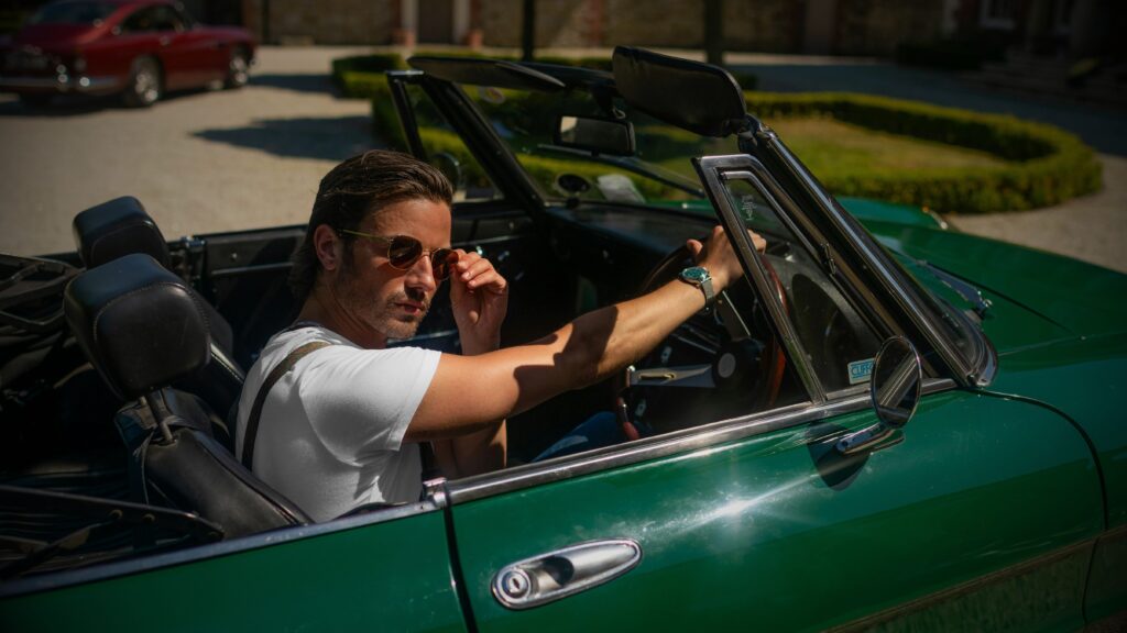 A man wearing a white t shirt, jeans, and sunglasses driving a classic Alfa Romeo Spider.