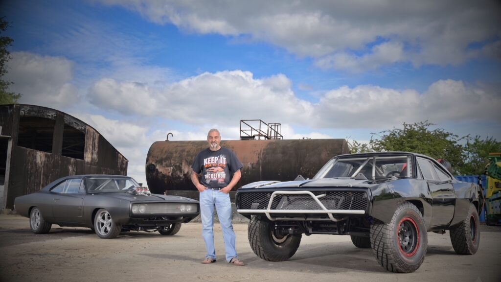 Greg McNair of Outlaw Muscle Cars standing between two custom classic Dodge Chargers: a matte black Fast and Furious 4 style replica and a lifted, rugged off-road Furious 7 movie car.