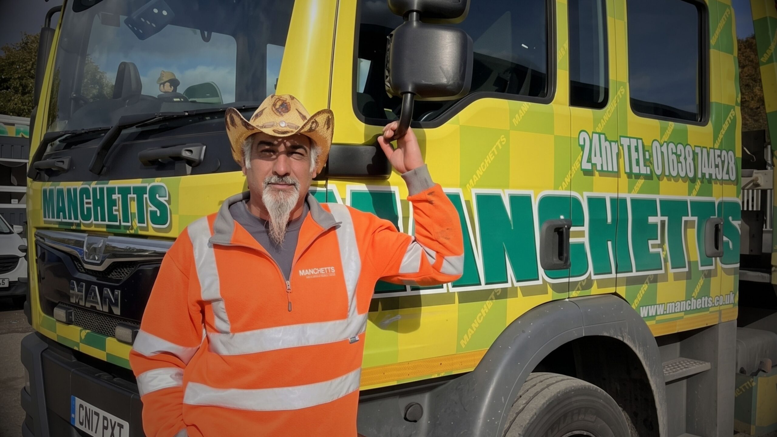 Greg McNair wearing a cowboy hat and high-visibility jacket, leaning against a yellow and green Manchetts MAN vehicle recovery truck.