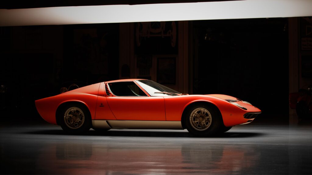 Alt Text: > Side profile of a classic 1960s Lamborghini Miura in vibrant orange with silver side skirts, parked in a dimly lit enthusiast garage under dramatic studio lighting.