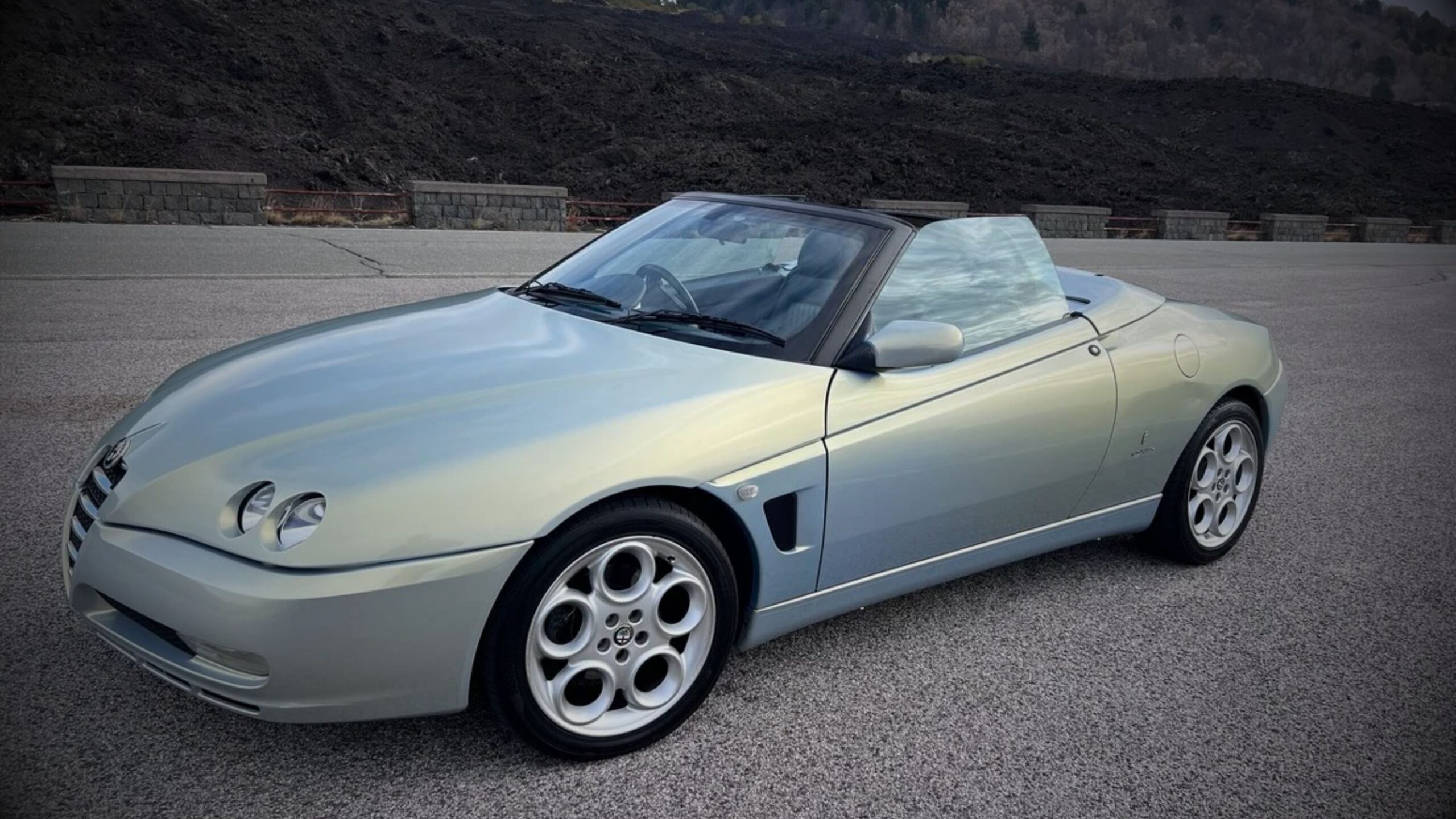 A silver or light iridescent blue Alfa Romeo Spider 916 convertible parked on an asphalt lot, featuring its signature twin round headlights and teledial alloy wheels against a dark, volcanic rocky hill background.