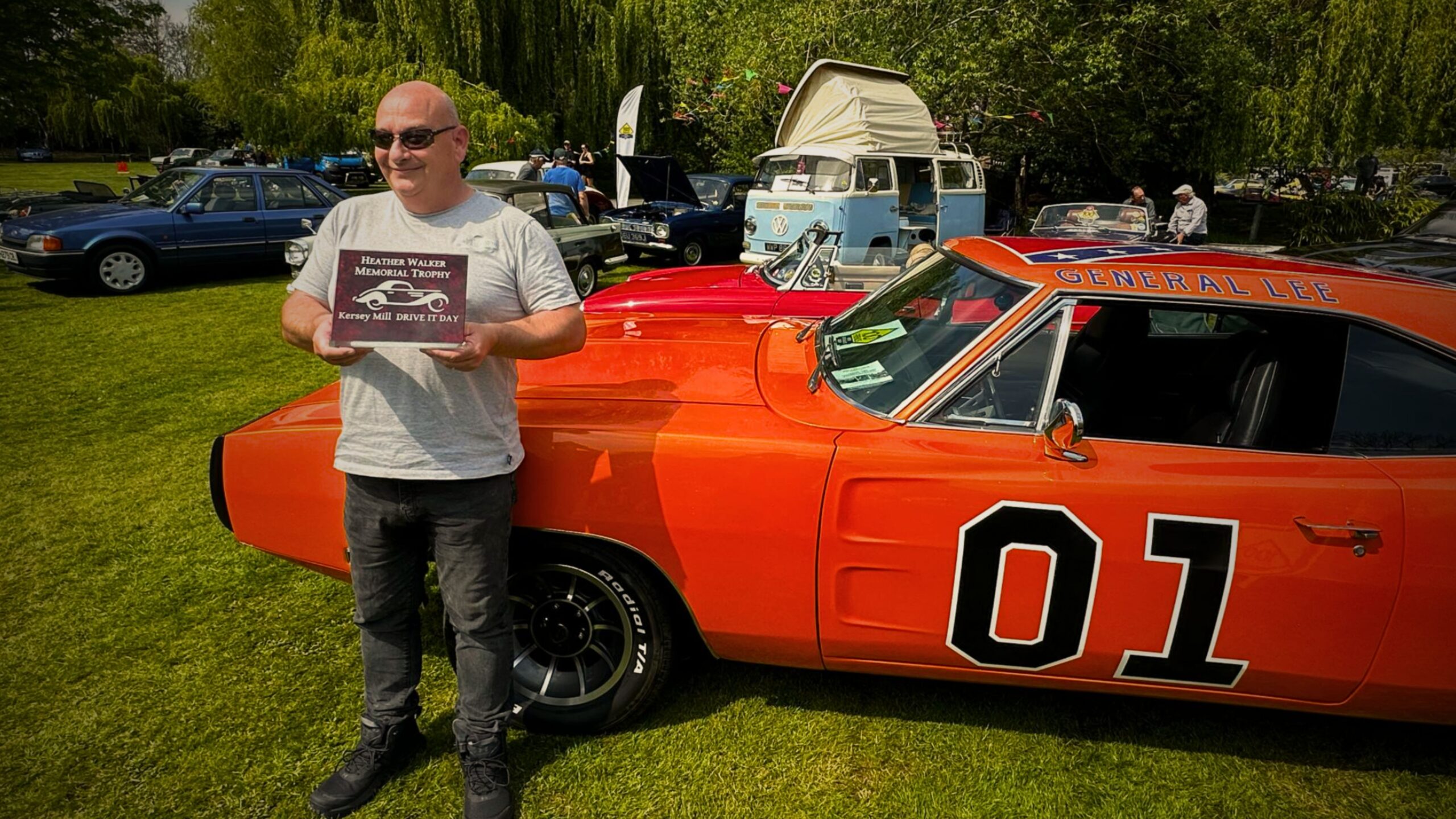 David Lay in sunglasses holding a "Heather Walker Memorial Trophy" plaque while standing next to a bright orange Dodge Charger "General Lee" recreation at an outdoor classic car show.