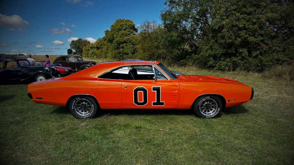 Side profile view of a pristine, bright orange classic Dodge Charger "General Lee" recreation parked on a grassy field on a sunny day.