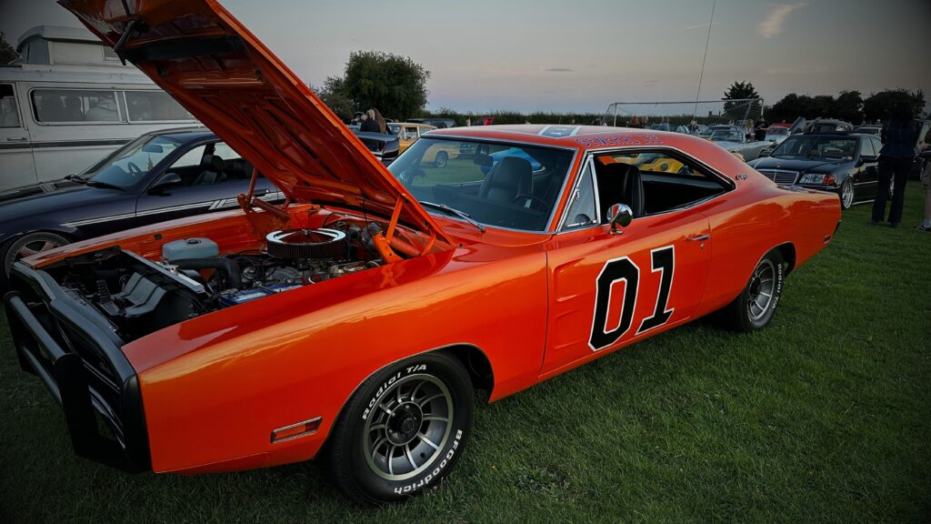 A three-quarter front angle shot of a bright orange Dodge Charger "General Lee" recreation with its bonnet propped open to display the engine bay at an evening car show.