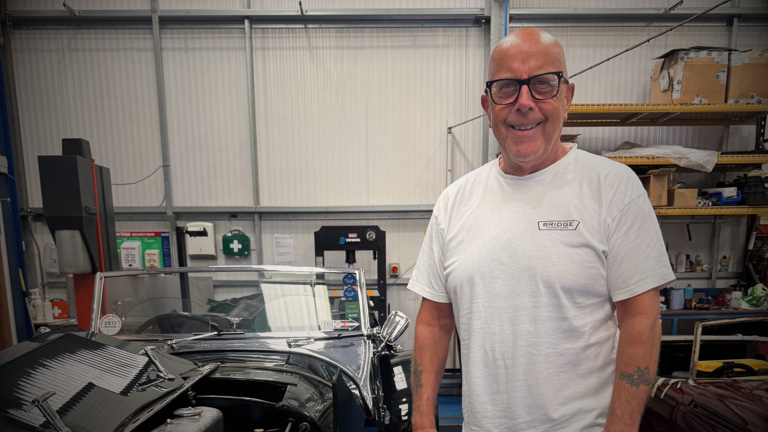 Founder and Director Gordon Ranson posing inside the Bridge Classic Cars workshop, surrounded by vintage vehicles under restoration