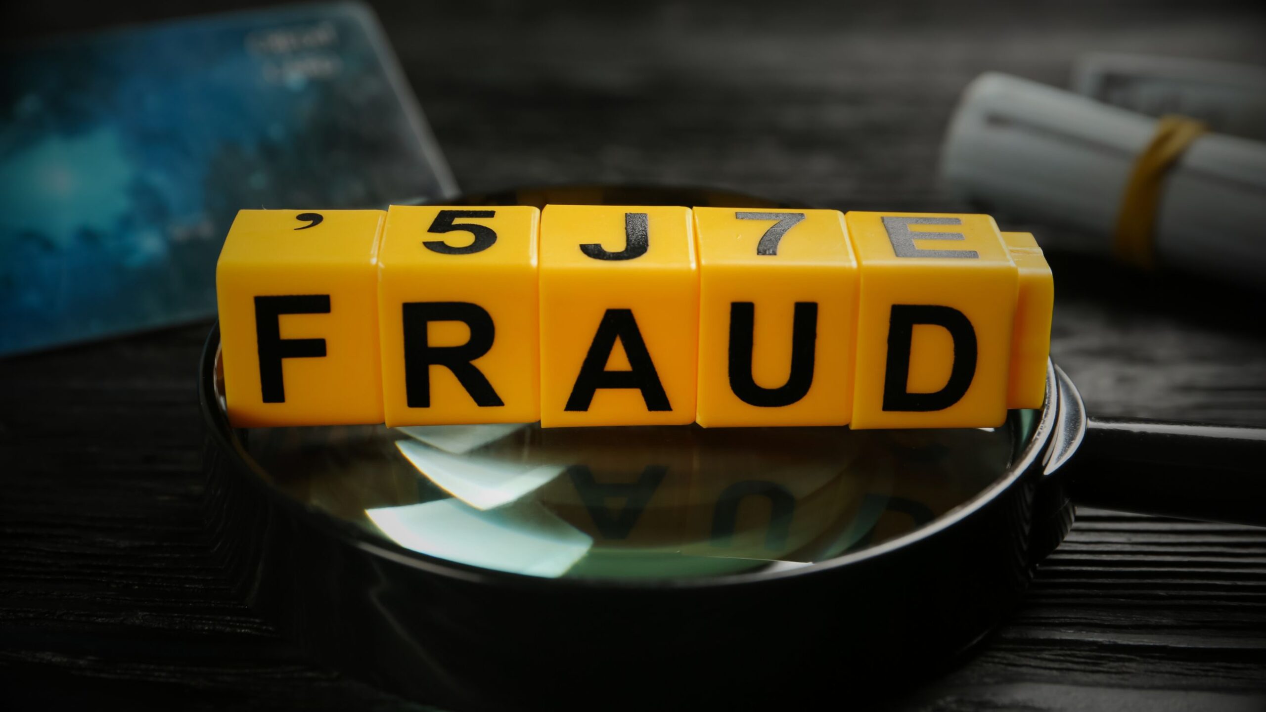 Five yellow toy blocks spelling out the word "FRAUD" sit on top of a magnifying glass lens against a dark background, with the word "INSURANCE" written in white text directly above them.