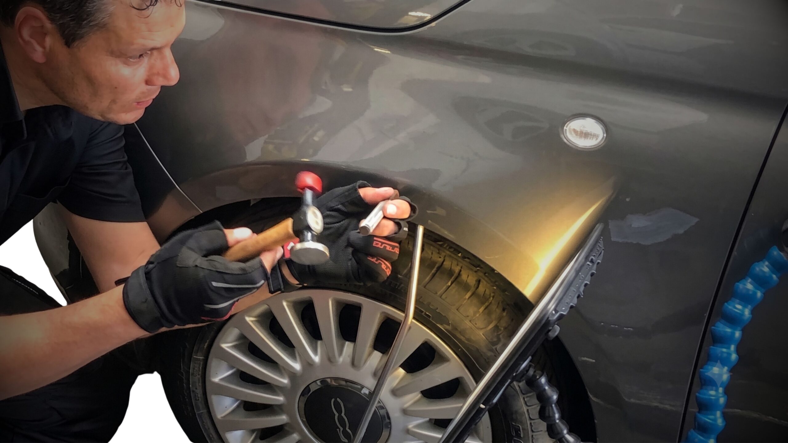 DENT REPAIRS 3 A close-up photograph of a paint technician crouching and meticulously performing a dent removal repair on the front fender of a grey car. He is wearing fingerless gloves and using a specialised hammer and punch tool, with a reflection board and blue flexible hose in the background.