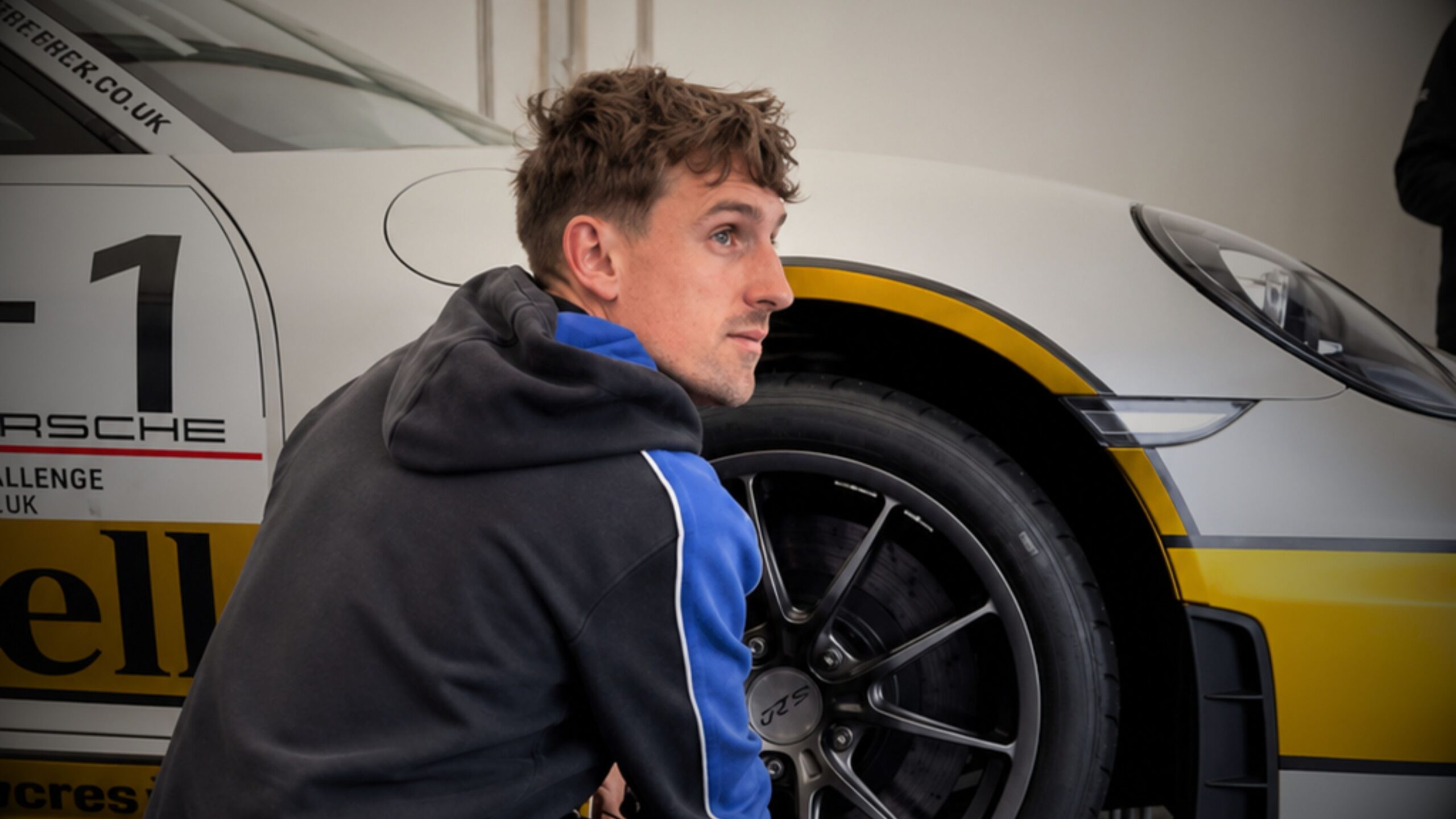 A photograph featuring James Barron, the owner of Autowerx, captured in a candid profile as he crouches near the front wheel of a Porsche 911 GT3 race car. The car is mostly white with a yellow stripe along the front fender. Part of the livery on the door is visible, showing the number "1" and the word "PORSCHE." James is wearing a dark charcoal hoodie with blue stripes on the sleeves and is looking towards the right side of the frame.