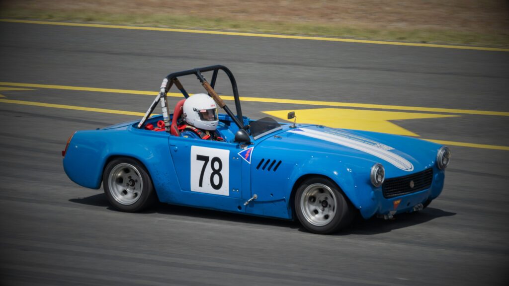 A classic blue MG Midget racing car with the number 78, a black roll cage, and white stripes, driving on a asphalt racetrack.