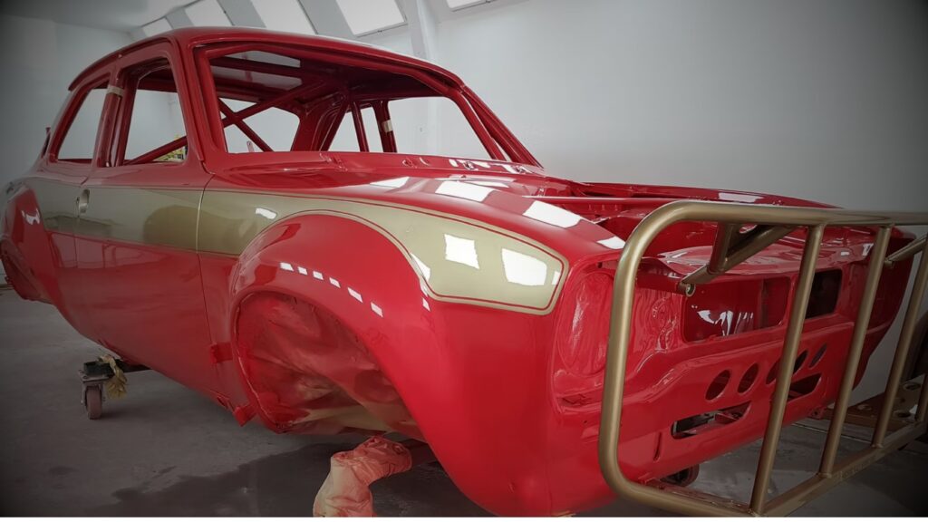 A fully painted red Ford Escort Mark 1 car bodyshell mounted on a rolling workshop cart inside a paint booth, featuring classic gold side stripes, flared wheel arches, and a tubular gold front tubular bumper guard.