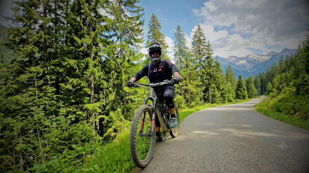 Mountain biker, Gavin Pink, full-face helmeted and fully geared up, pauses on a paved mountain path in the French Alps surrounded by dense pine trees with snow-capped alpine peaks in the background.