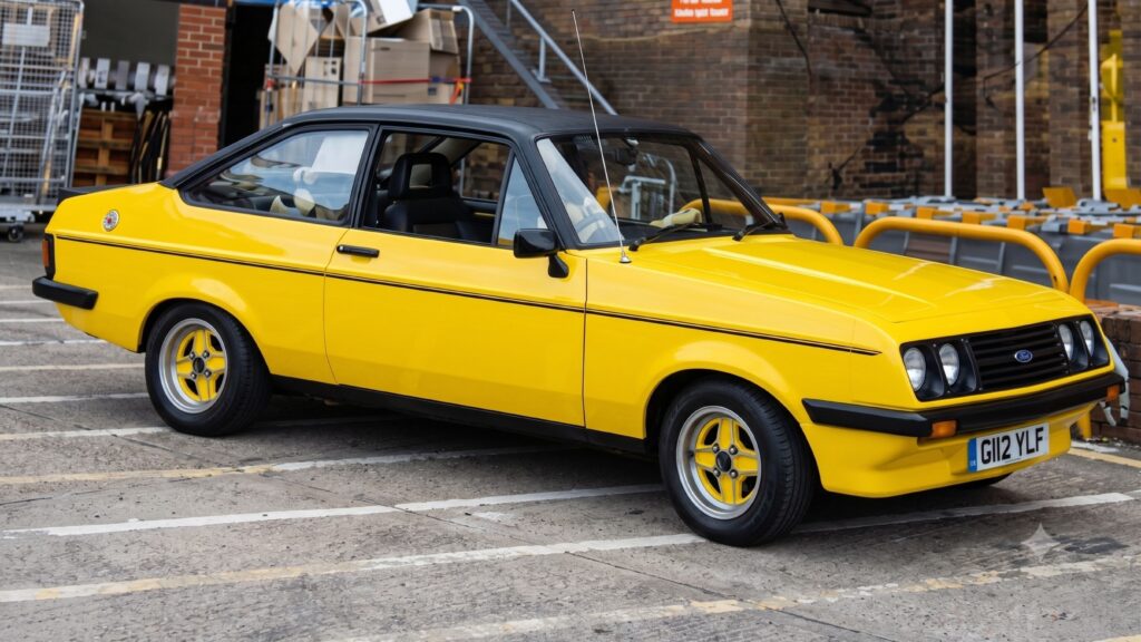 A bright Signal Yellow Ford Escort RS2000 Mk2 classic sports saloon parked on a paved lot, featuring a black vinyl roof, slant-nose front grille, black side pinstripes, and colour-matched four-spoke RS alloy wheels.